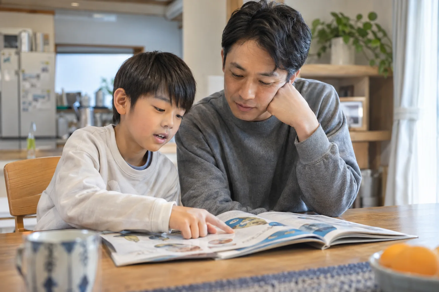 開いた本を指さす子どもの話を、家庭で父親が聞く様子